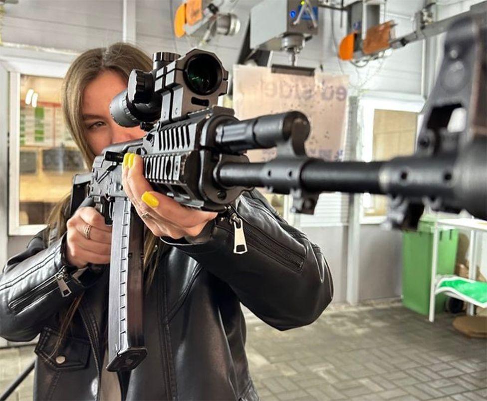 A young woman with long brown hair poses with an automatic rifle in what could be a shooting range. She is looking through the telescopic lens, with the muzzle of the gun facing the camera. She has bright yellow nail varnish and wears a leather jacket. 