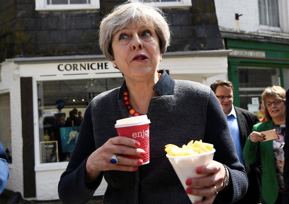 Theresa May holding a hot drink and some chips