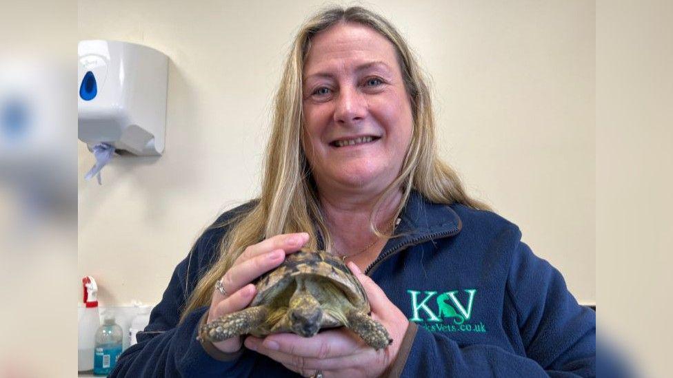 Sara Marchant holding the tortoise and smiling into the camera. She has long blonde hair and is wearing a blue fleece with the Kirk Vets logo on it.