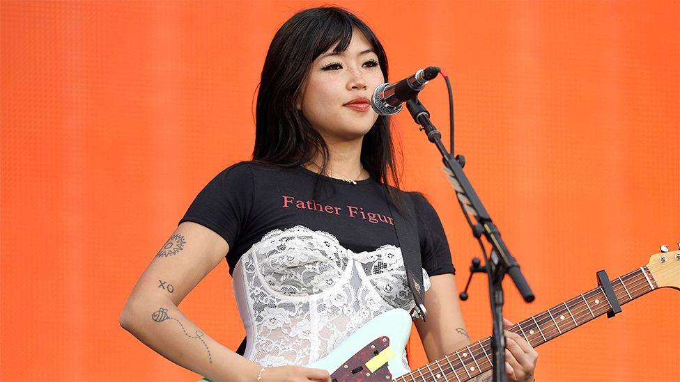 Beabadoobee playing a guitar at a concert with an orange background