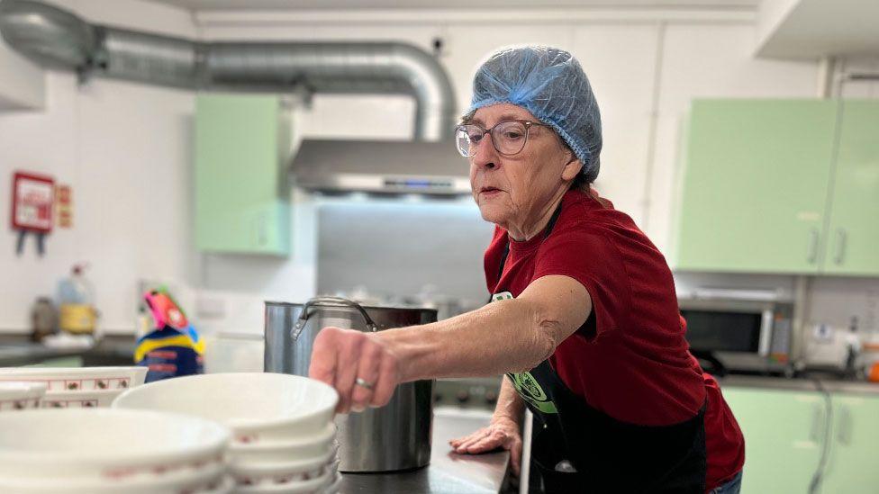 Maggie Bointon, wearing a blue food-safety net over her hair, glasses and a red T-shirt. She is in a kitchen and is leaning over a stainless steel counter, reaching towards a pile of bowls. In front of her is stainless style pot from which steam is rising.