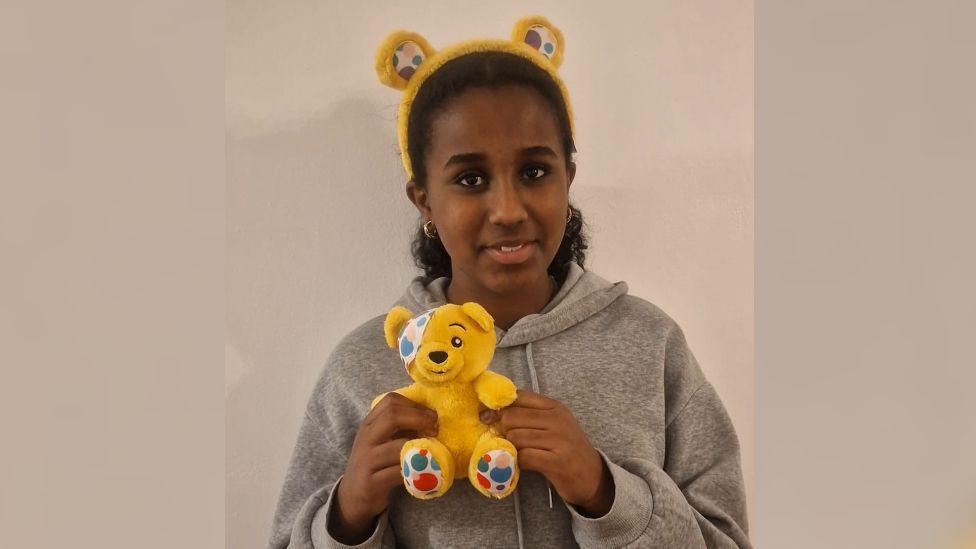 Bethel smiling holding a yellow Pudsey bear with yellow Pudsey bear headband. She is wearing grey hoodie. 