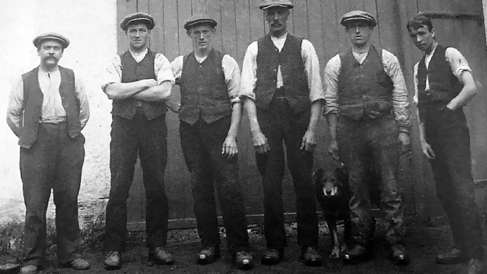 A black and white image of a row of distillery workers in waistcoast and bunnets with a dog