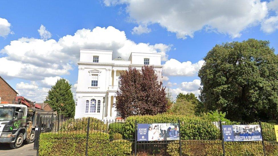 Google StreetView image of the white historic school building behind trees and hedging.