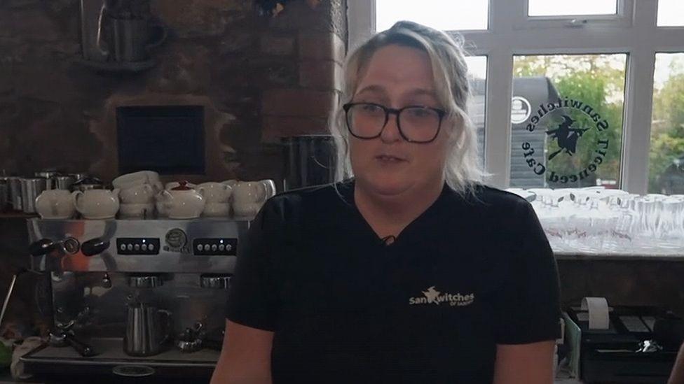 Cafe owner Christine Dobson Moore standing in front of a window in her cafe. She has blond hair tied back and is wearing dark framed glasses and a black tshirt. There are cups on a coffee machine to her left