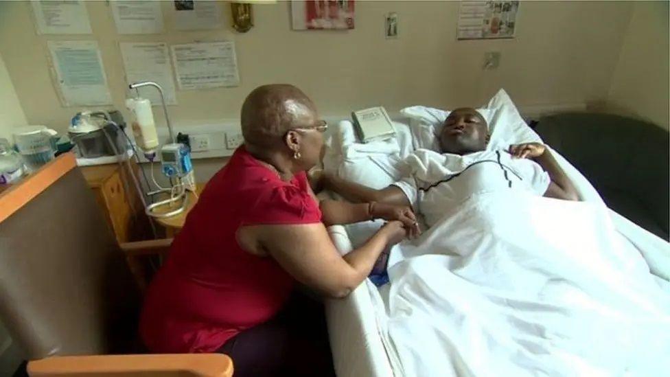 Julian Cole in a bed. A woman in a red top sits on a chair next to it, with her forearms resting on the side of the bed. There is some sort of drip near the bed.