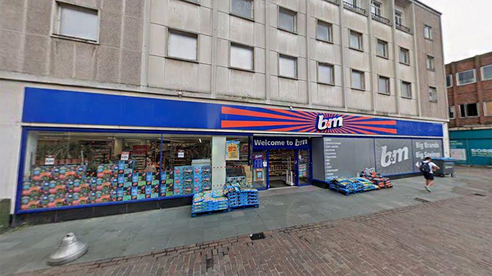 The outside of B&M in Bedford, with a man walking past the store. Above the store front is a big bright B&M sign in blue and orange, and there are products stacked in the window.