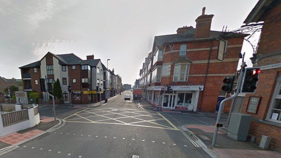 Four-way road junction with yellow hatchings on road in centre and buildings on all four corners it is a bright and dry day.