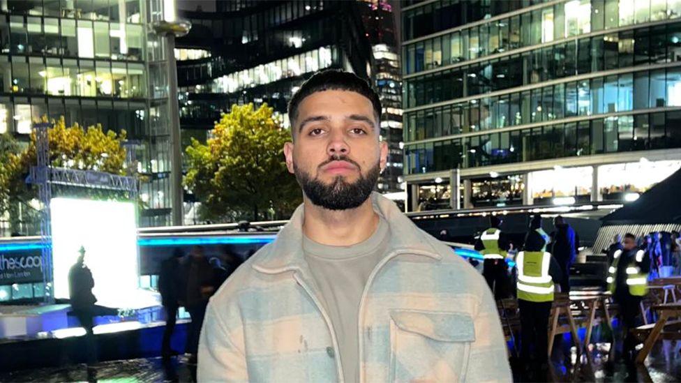 Vaibhav, a male, wearing a light blue and pink jacket, with the lights of buildings behind him.