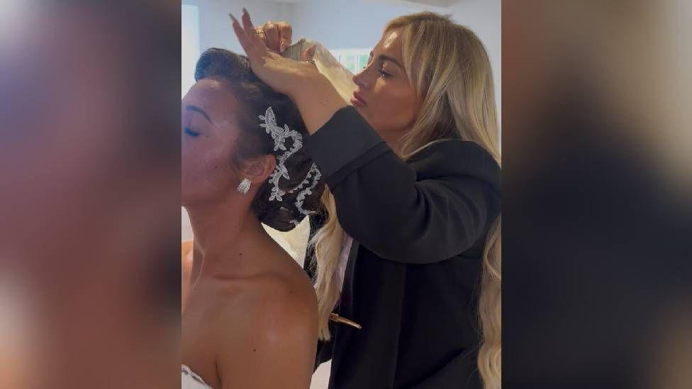 A blonde woman styling the hair of a woman in a wedding dress. the woman in a wedding dress has some jewellery in her hair.