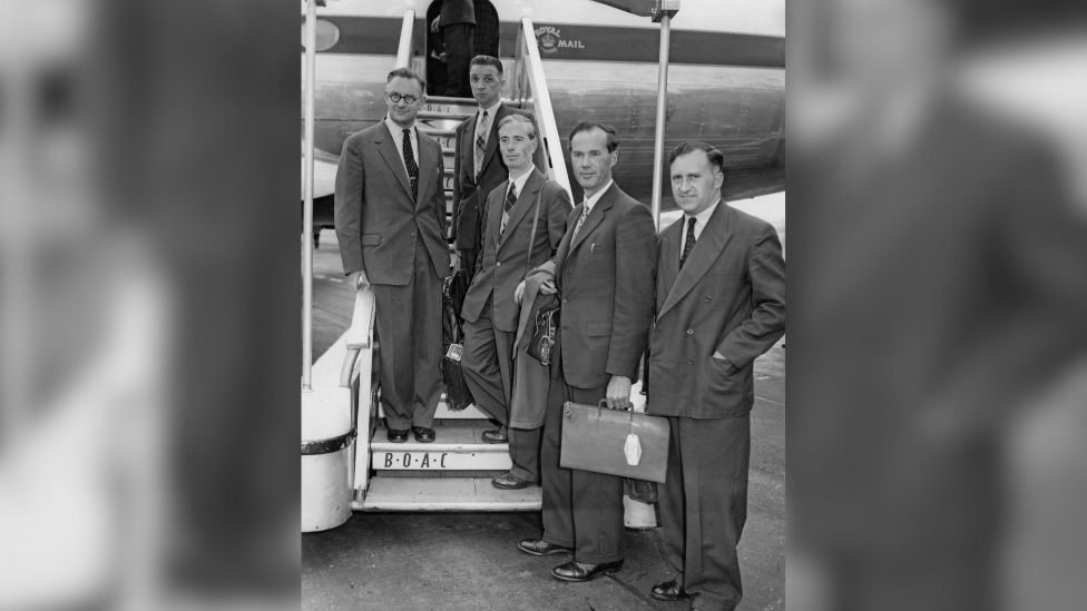 Black and white image of James Sayers (second from right) leaving London Airport with other scientists. James is carrying a bag. The three others beside him are standing on the steps of the plane