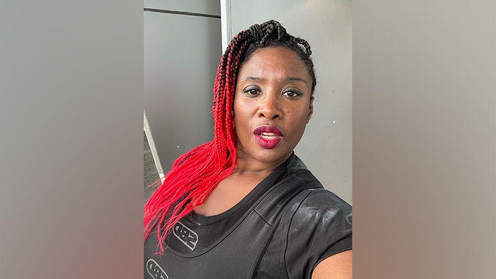 Selfie head and shoulders shot of Elaine Jackson with red braided hair tied in topknot pony tail and she's wearing a black t-shirt and black vest on the top.