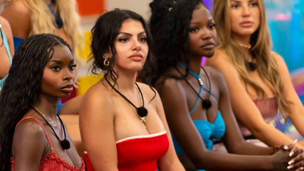 From left to right sitting in a row: Love Island USA contestants Olandria Carthen in a strappy red top, Huda Mustafa in a red strapless top with white ribbing, Michelle "Chelley" Bissainthe in a blue strappy top and Iris Kendall in a dusky pink strapless top.