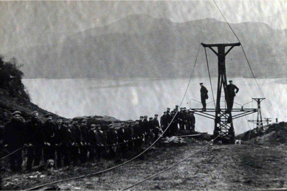 Archive photo of a workers lined up alongside a pulley system for taking materials from the Inversnaid area of Loch Lomond to Loch Arklet, where a dam was built as part of the Katrine Aqueduct project