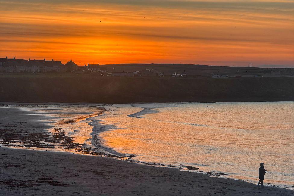 Lone silhouetted figure on a beach, with an orange sunrise sky overhead, reflected in the sea.