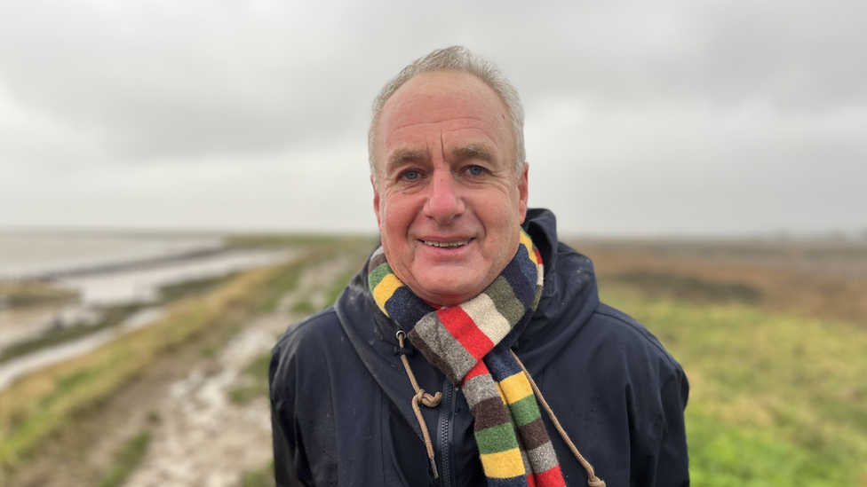 Man with short white hair wearing a scarf with coloured stripes and a raincoat