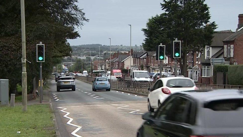 A view of a road with cars driving down two lanes. Houses line the right of the road and grass and trees are on the left hand side of the road.