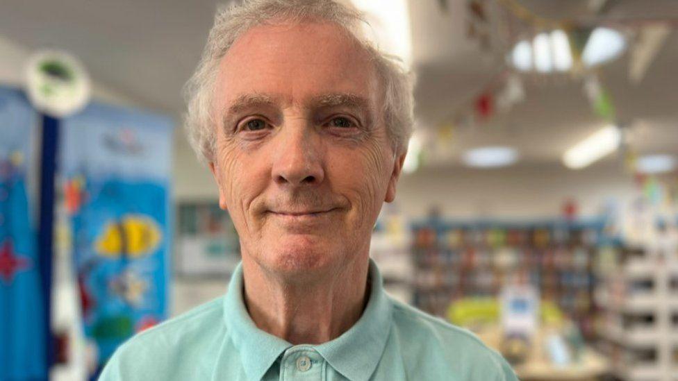 Smiling man with short grey hair and peppermint coloured polo shirt - shelves of books can been seen blurred in the background