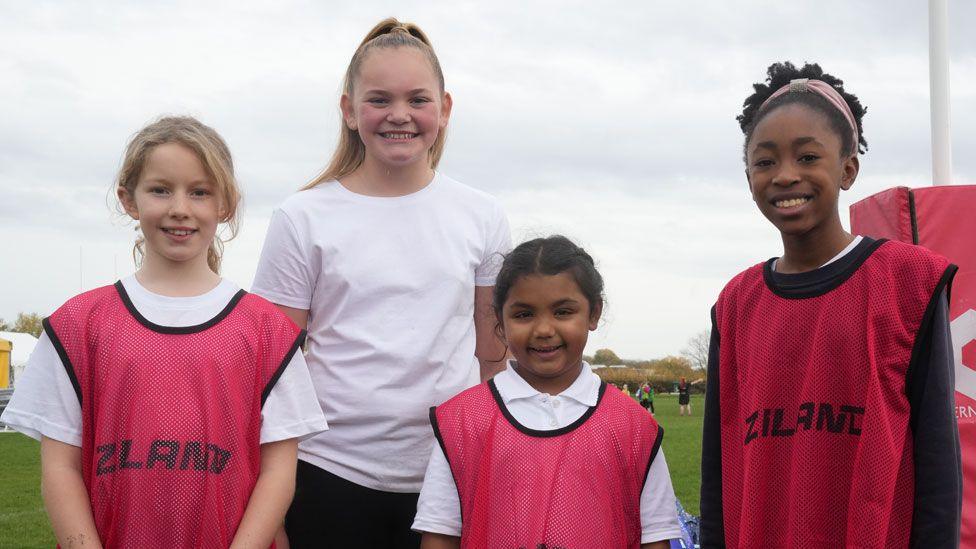 Four primary school girls in a row. The three in front are wearing deep pink tabards over white T-shirts and the one behind is wearing a white T-shirt. They are all smiling broadly and the rugby pitch can be glimpsed behind.