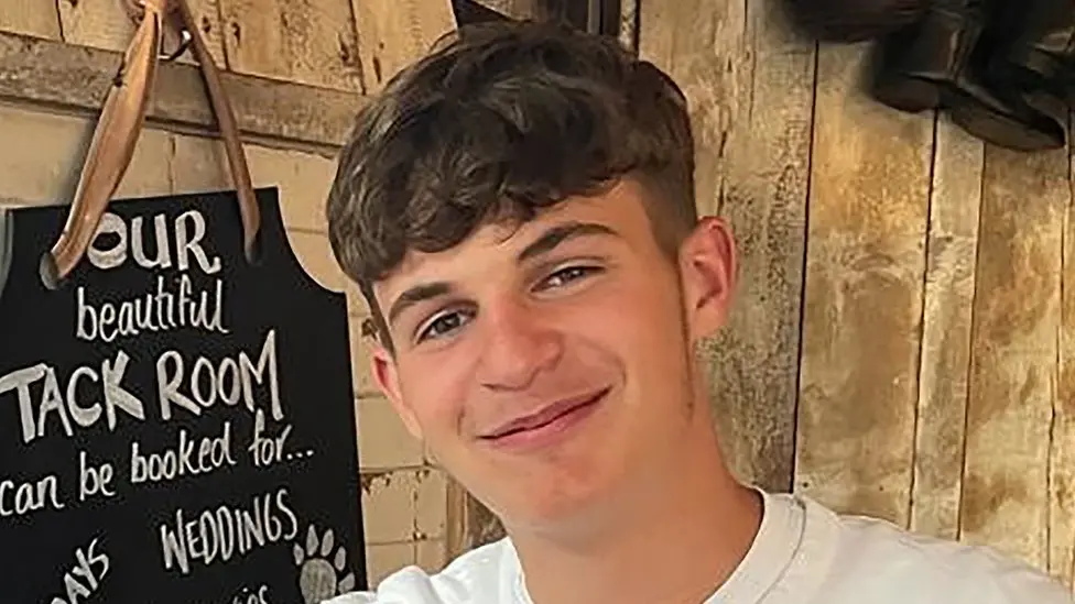 A photo of Billy Lardner smiling at the camera. He has short, brown hair and seems to be sat inside a wood-panelled room or shed 