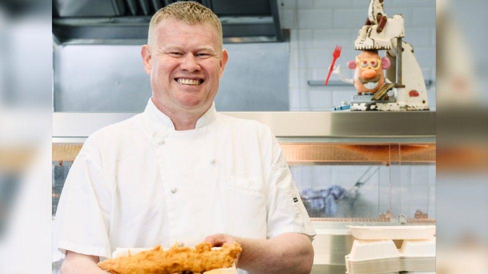 Andrew Crook CFSP, President of the National Federation of Fish Friers facing the camera with a batted fish on a plate in his hands.