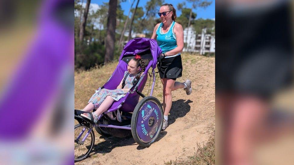 Woman with brown hair running with her daughter who is sat in a wheelchair