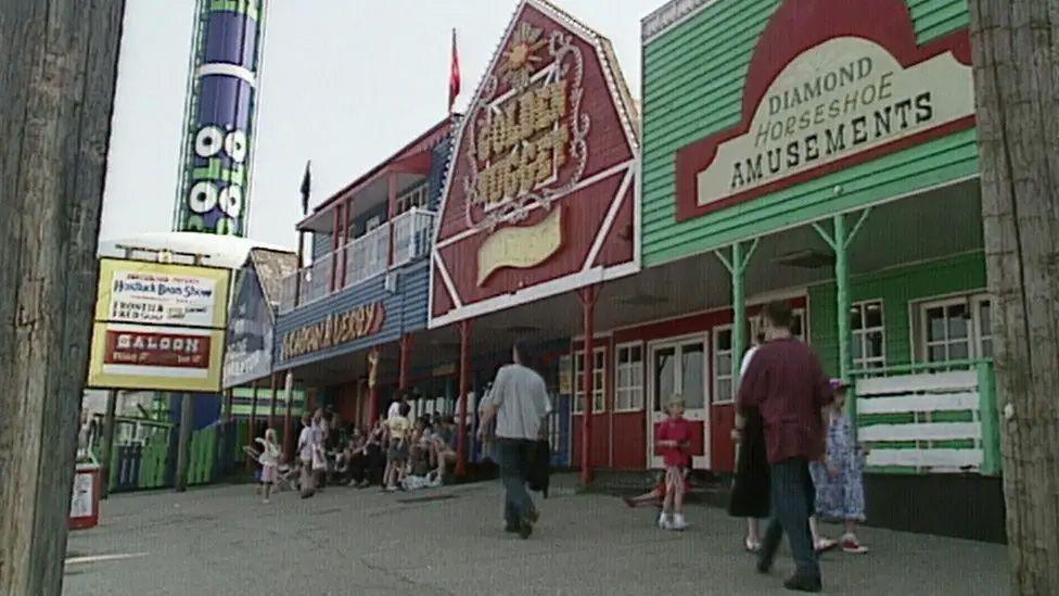 A row of shops at the former Frontierland theme park, in red and green, one saying Diamond Horseshoe Amusements, one saying Golden Nugget. Peole are strolling past and in the background is a ride that looks like a giant tube of polo mints