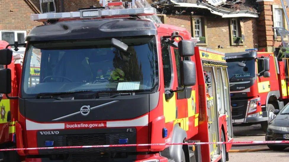 Red and yellow fire engine with "Bucksfire.gov.uk" in white lettering on the front. There is another fire engine just visible to the left and a third behind. A two-storey building with a collapsed fire-damaged roof is visible behind.