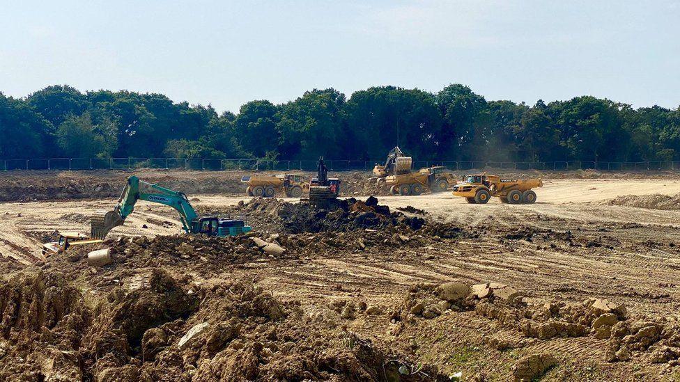 Diggers churn up a large muddy plot of land on a sunny day.