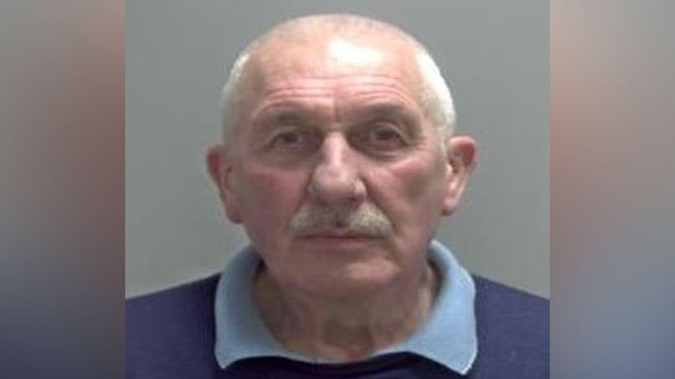 A police custody shot of Alan Johnson with a white moustache and hair. 
He is wearing a navy blue jumper with a light blue polo shirt underneath. 