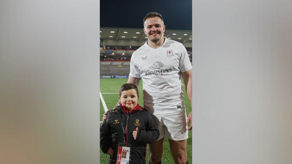 Ollie gives the thumbs up beside Jacob Stockdale on the pitch. Stockdale has black hair and facial hair and is wearing the white Ulster kit and shorts.