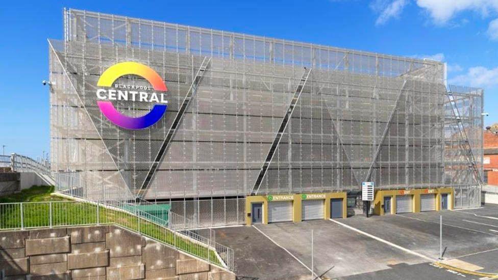 Street view of the central multi-storey car park, a large grey framed building with a rainbow coloured circular sign on the front left saying 'Blackpool central'