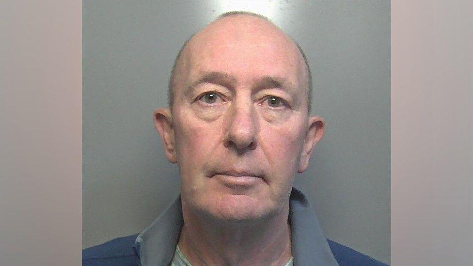 A custody photograph shows Graham Tomlinson looking directly at the camera. He has close cropped hair and is wearing a blue and grey fleece/