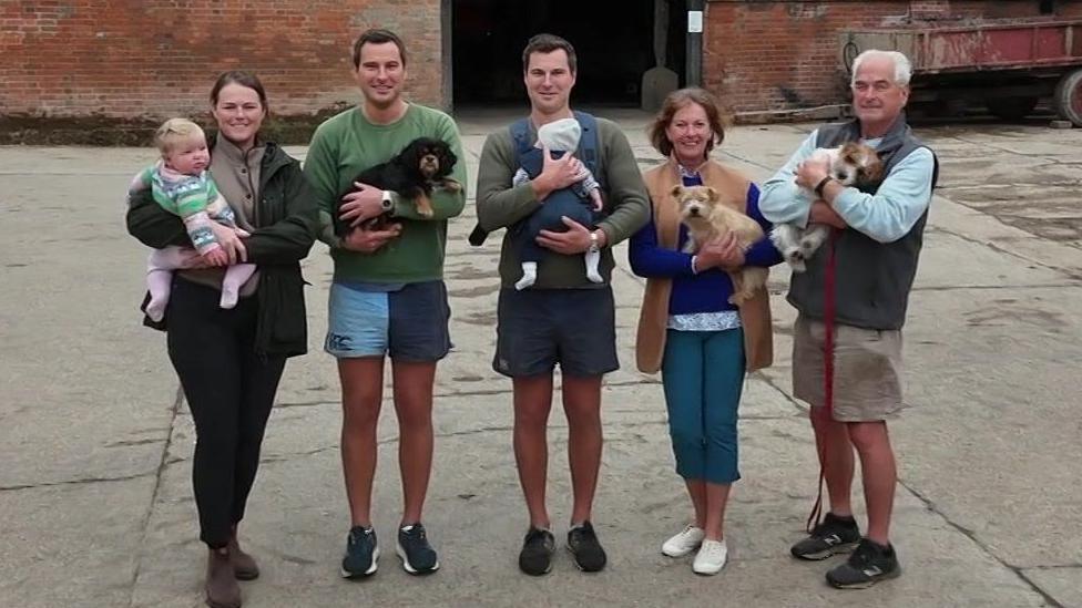 From the left in a line stands Georgie, Tom, Will, Sandra and Peter Carlisle. They are standing in front of their farm buildings on a concrete yard. Georgie is holding her baby daughter and wearing a green coat and black trousers. Tom is holding his black spaniel, wearing shorts and a green jumper. Will is holding his baby in a front carrier and is wearing shorts and a green jumper. Sandra is holding a small dog as is Peter.