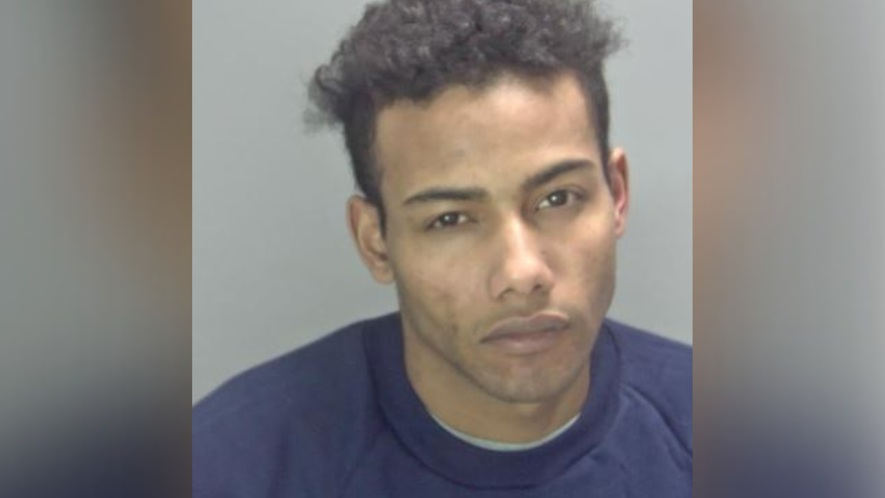 Police custody photo of Junior Suleimane against a grey background. He is wearing a plain blue crew neck jumper, has short brown curly hair and brown eyes.