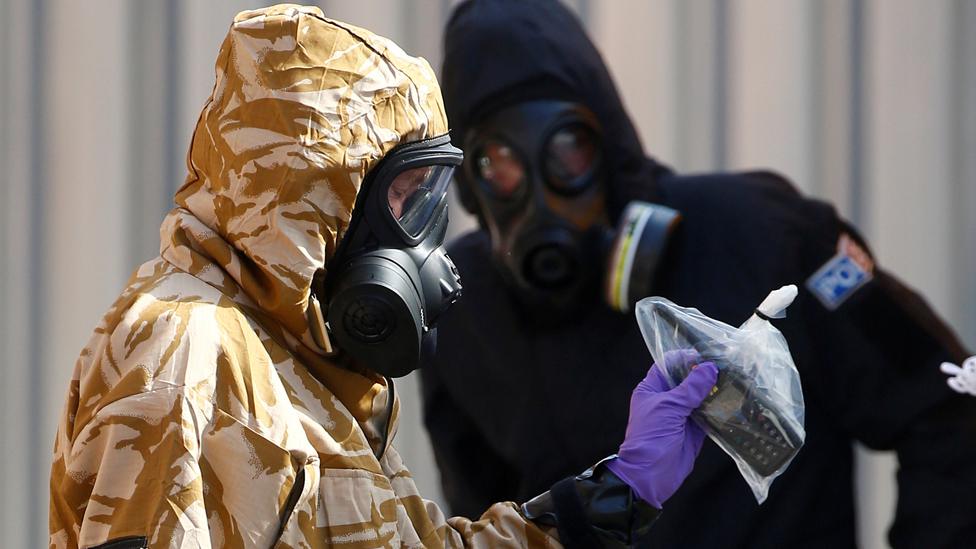 Forensic investigators, wearing protective suits, emerge from the rear of John Baker House