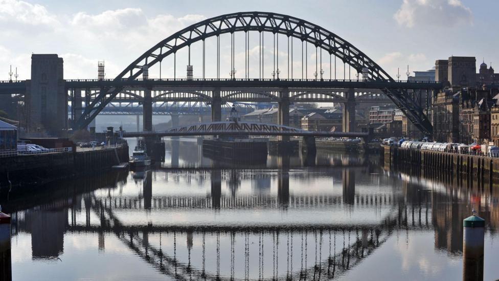 The Tyne Bridge, that stretches over the River Tyne. Pictured on a clear day with the bridge reflected in the river below. Other bridges such as the Swing Bridge and High Level Bridge can be seen in the background.