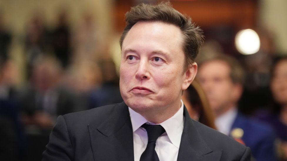 Elon Musk, with dark hair wearing a black suit and tie with a white shirt, looks to his right while sat in an audience with blurred out people behind him 