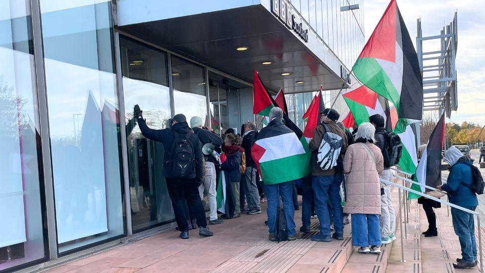 A group of people holding red, white, green and black Palestine flags crowd around the glass door of a BBC Scotland building.