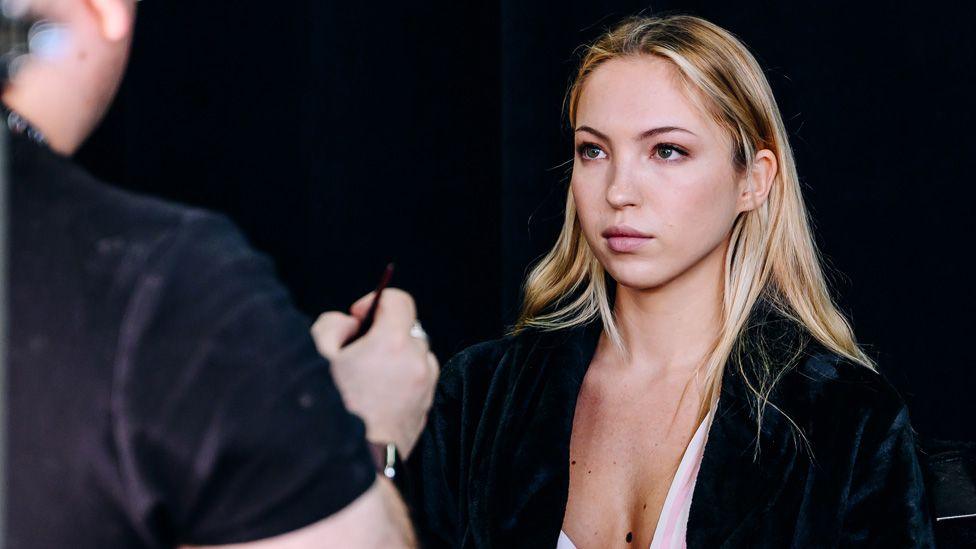 Lila Moss sitting backstage as a figure appears to prepare to apply make-up 