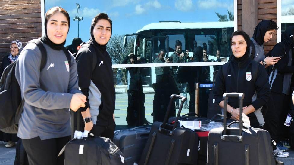 Women in dark clothing stand with their luggage. Behind them is the reflection of a bus in a glass window