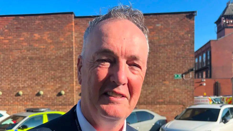 Close up of Clive Grunshaw smiling, standing in a car park with a large red brick building in the background and a stripe of blue sky above it