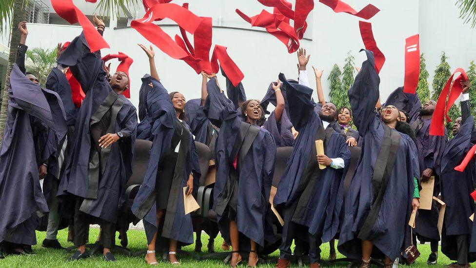 Cheering business graduates in black robes, standing on green grass, throw red scarves into the air in Lagos, Nigeria - 2019