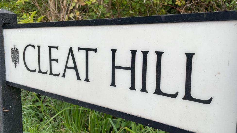 A close-up of a white road sign saying Cleat Hill in black block capitals. Behind it is grass and foliage.