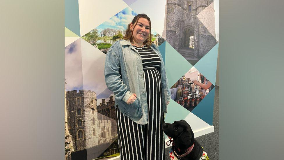 Molly Watt - with long brown hair, tied back, with a dark blue and white dress and a denim jacket standing outside the 91ȱ Radio Berkshire studios with Bella, a black labrador 