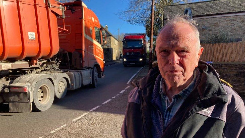 Man with very short hair wearing big grey coat stands next to stationary lorries on A422