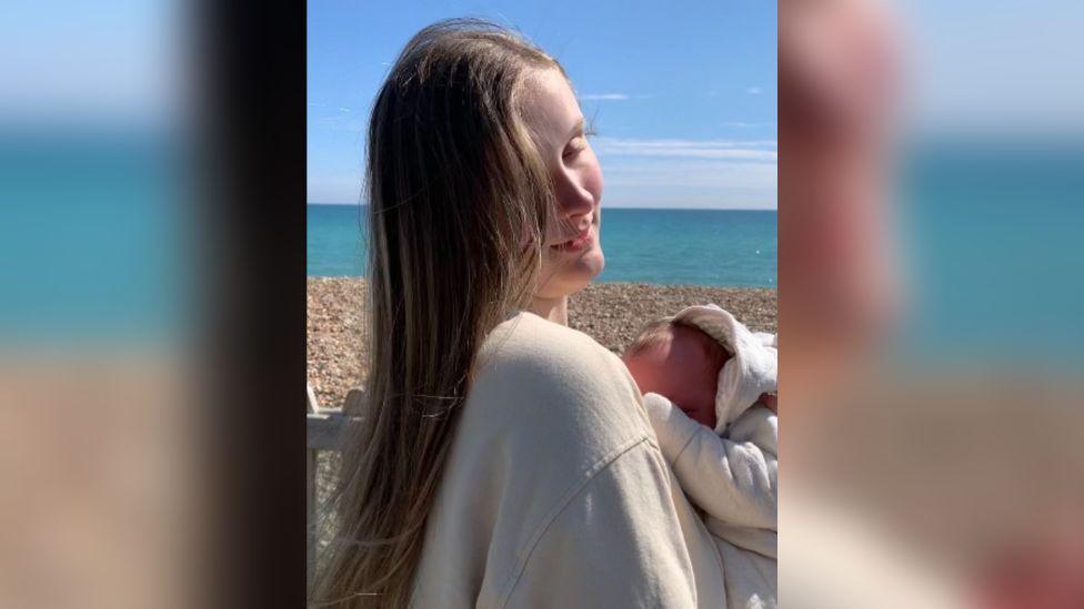 Morgan stands on a beach cradling her newborn baby. She has long blond hair and wears a white jumper. The baby wears a white babygrow. 