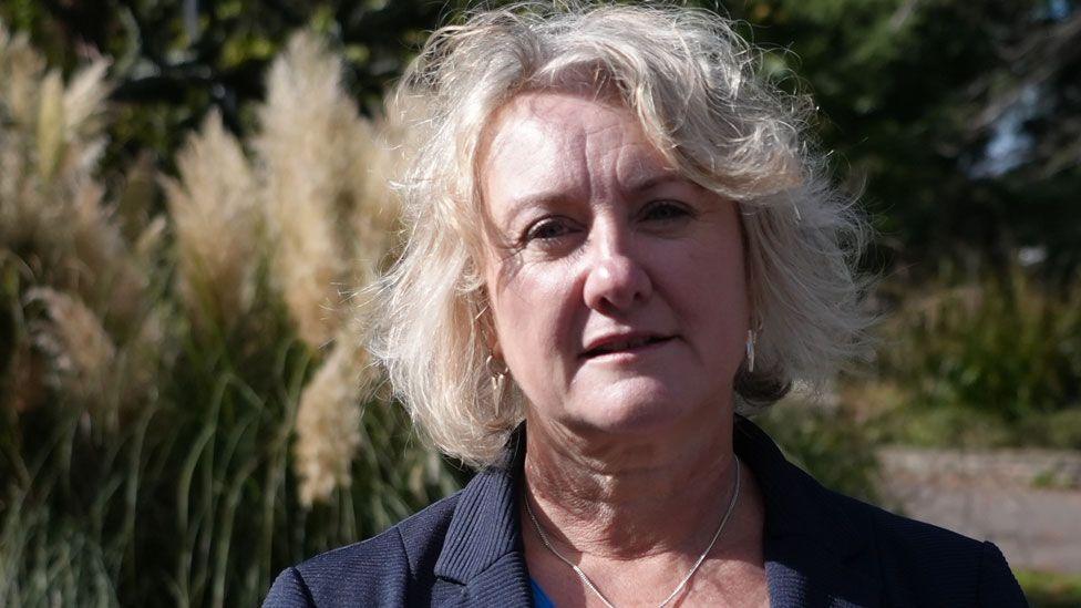 A close-up of Jo Procter, who has white hair in a bob and is wearing a navy blue jacket. She is standing outside with pampas grass and trees behind her. 