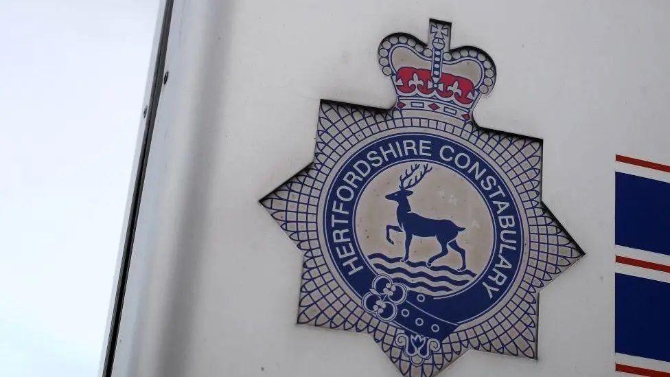 The emblem of Hertfordshire Police against a white sign. It is blue and white with a deer standing on a river.