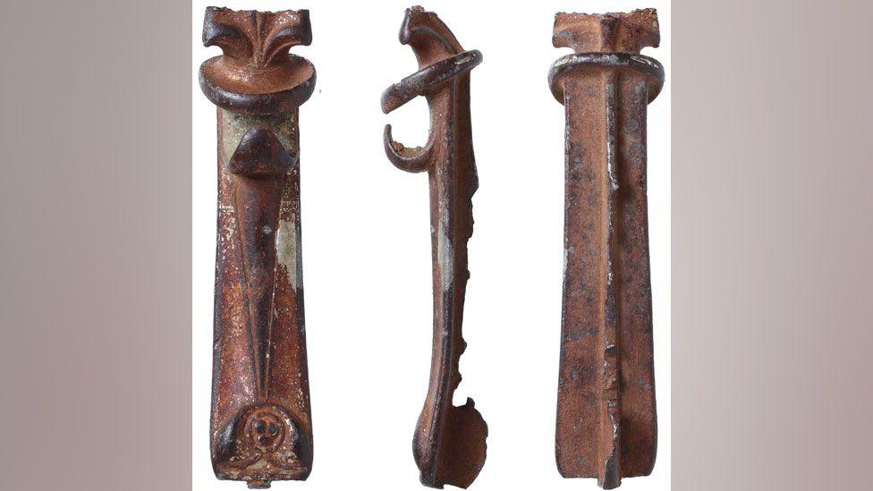Three views of a  long and slim copper alloy birdlip-type Iron Age brooch. On the left is shows its front, has mouldings along its body and a curved hook or beak at the top. The middle view is side on, showing the curving beak at the top and a ridge above it.  The right view is of the back which has ridge running along its length. It is missing its fastening and its top moulding has snapped off. 